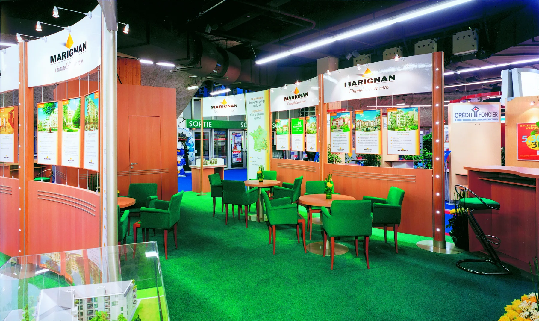 Le stand Marignan des salons de l'immobilier : fauteuils verts, tables rondes et panneaux aux cartes de France.
