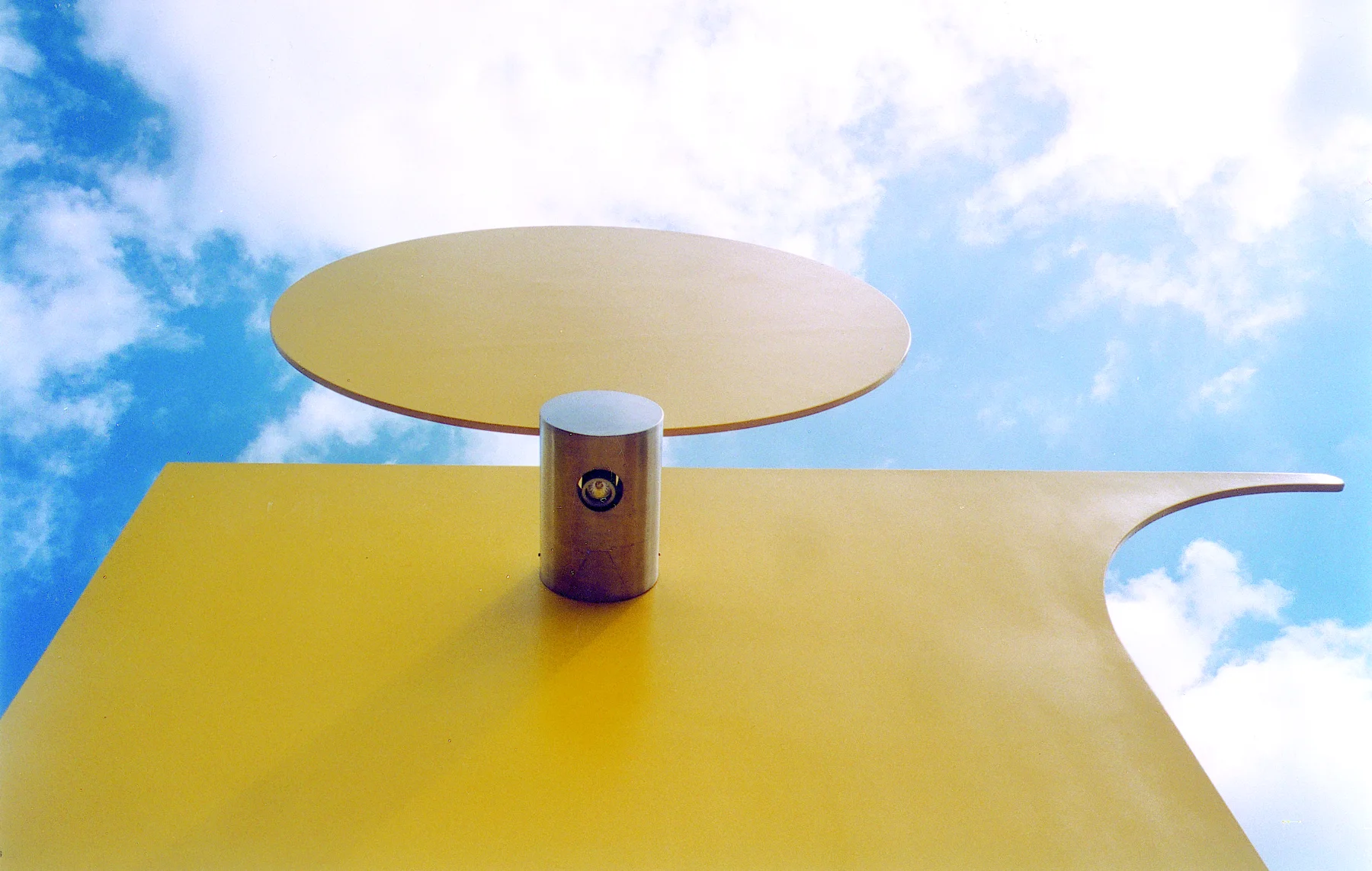 Le sommet du relais vu d'en bas : le point du « i », un disque jaune posé sur une pièce d'inox, contre le ciel.