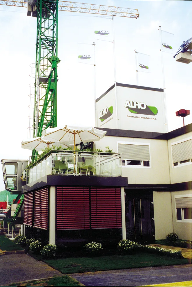 Stand ALHO construit en modules empilés, terrasse avec parasol et jardinières, logo ALHO en façade.