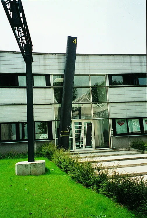 Mât noir incliné devant un immeuble de bureaux, sur une pelouse.