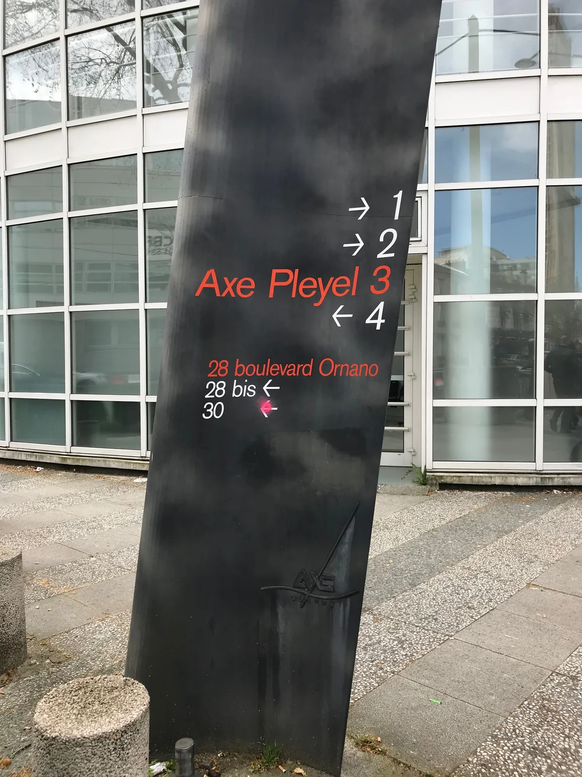 Totem noir incliné portant « Axe Pleyel 3 » et des flèches numérotées, devant un hall vitré.