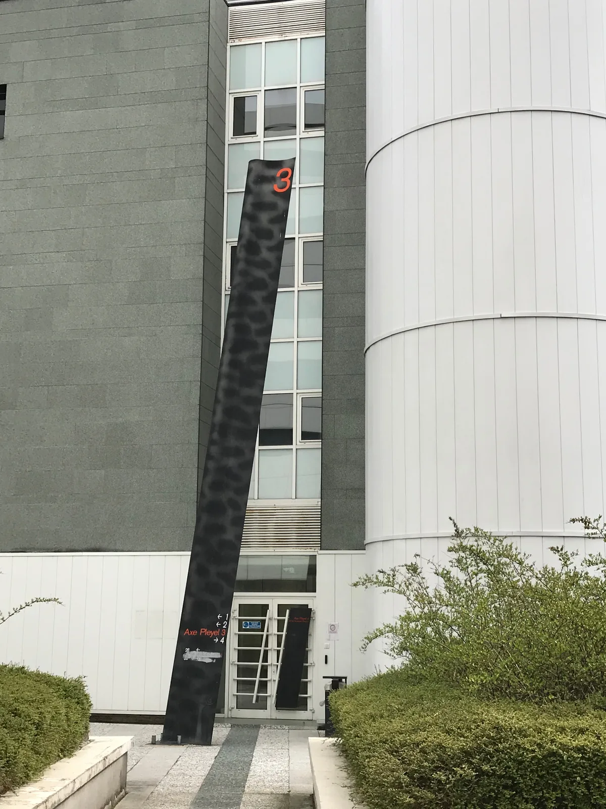 Haut totem noir dressé devant un immeuble gris et une tour ronde blanche.