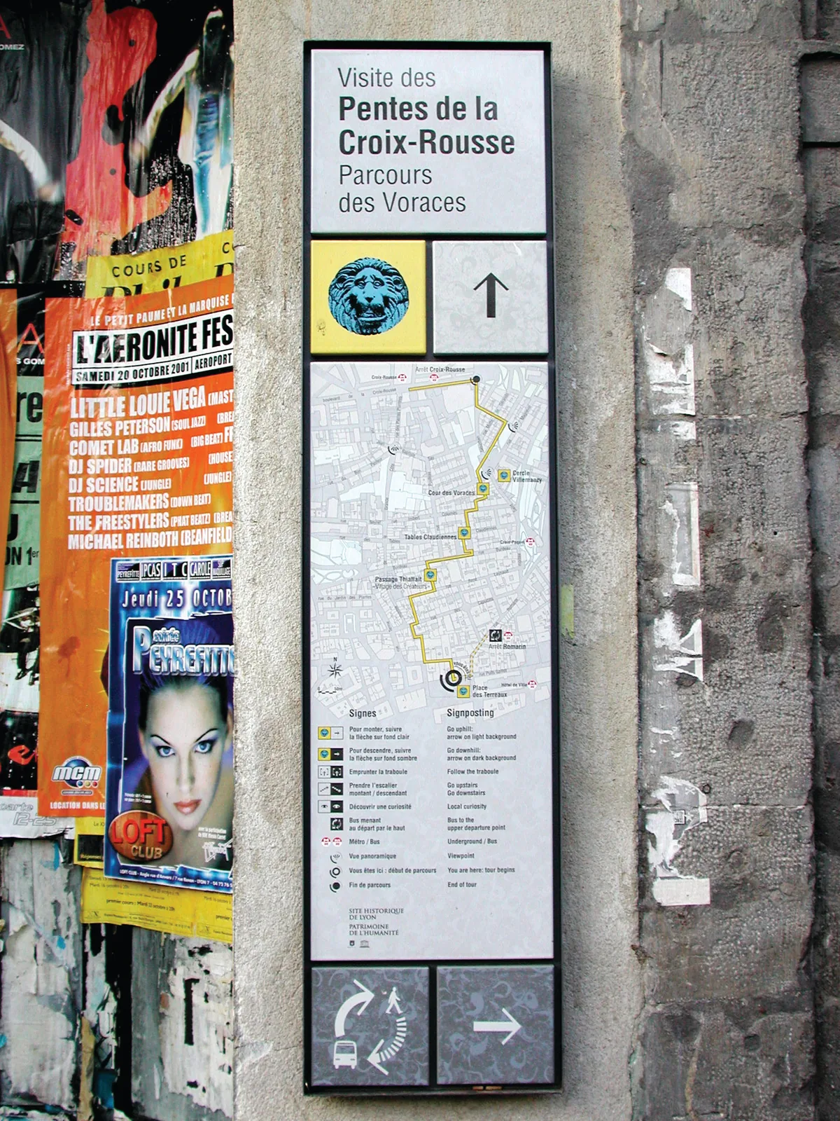 Panneau « Visite des Pentes de la Croix-Rousse, Parcours des Voraces » posé sur un mur couvert d'affiches.