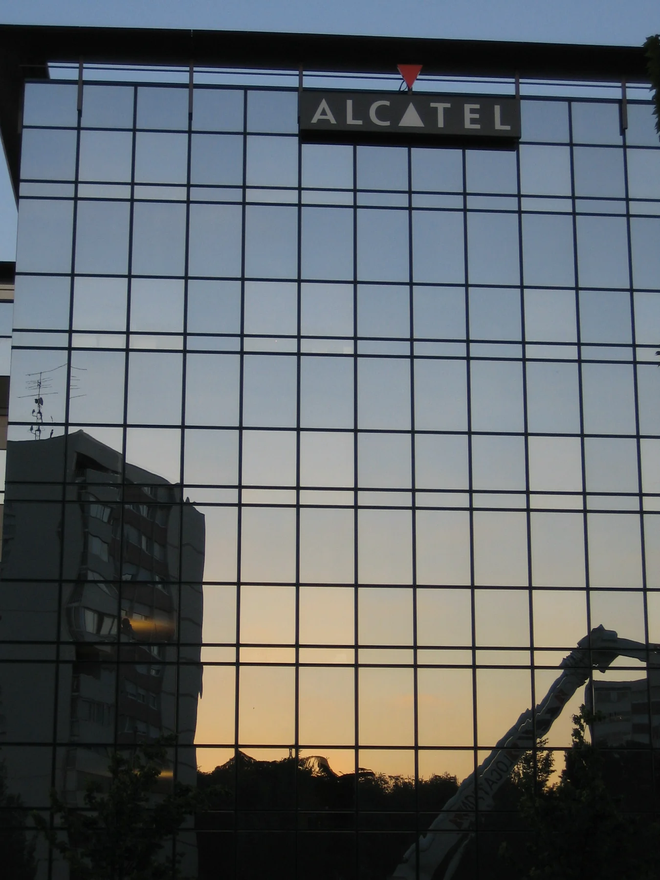Façade de verre au crépuscule, l'enseigne Alcatel allumée en haut du bâtiment, le ciel orangé reflété dans les vitres.