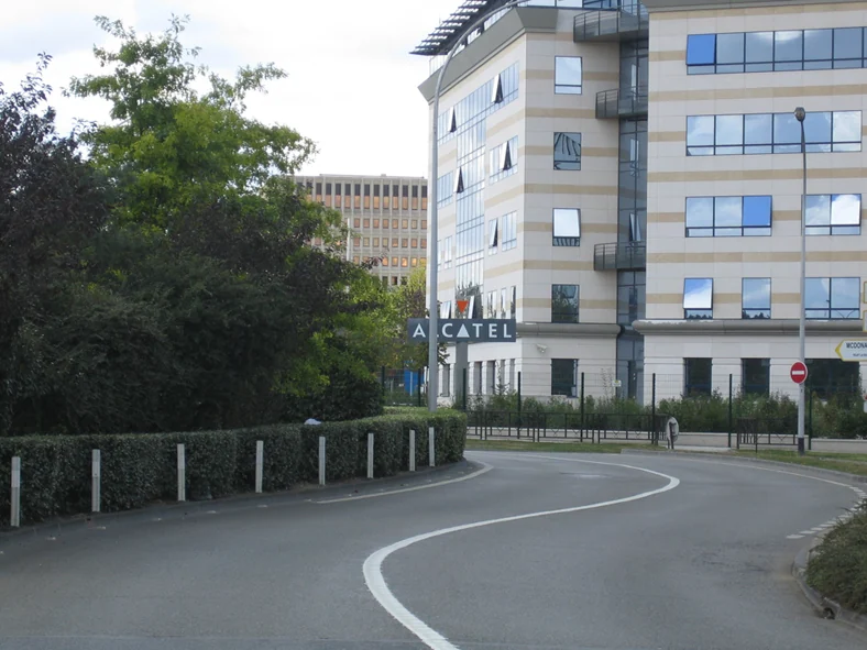 Allée courbe menant à un bâtiment de bureaux du site Nieuport, son enseigne Alcatel en façade, entre les arbres.