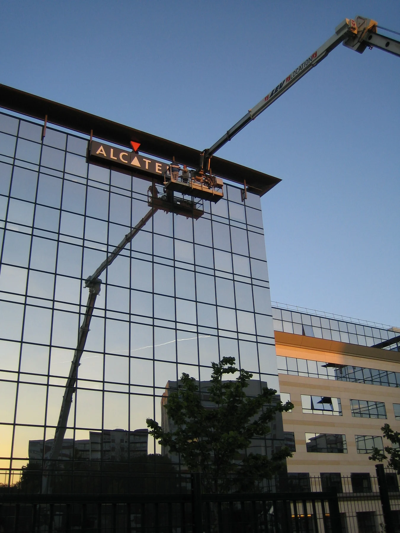 Une nacelle élévatrice devant une façade vitrée au soleil couchant : la pose de l'enseigne Alcatel en haut du bâtiment.