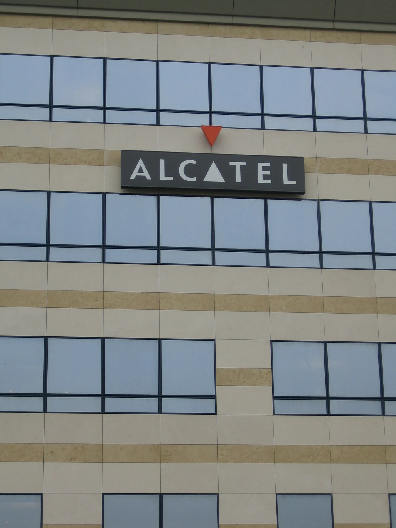 Enseigne Alcatel gris foncé posée en applique sur le parement de pierre claire, triangle orange au-dessus, entre deux rangs de fenêtres.