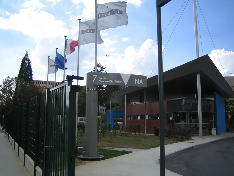 Poteau directionnel de l'entrée principale : bandeau cintré gris « 7 Avenue Morane-Saulnier, NA » devant les drapeaux du site.