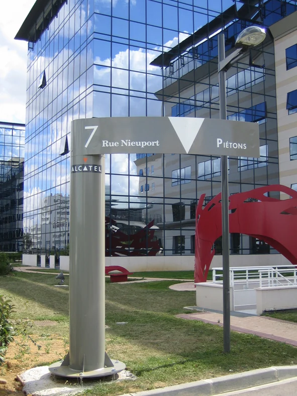 Poteau directionnel « 7 Rue Nieuport, Piétons » sur la pelouse devant un immeuble de verre, une sculpture rouge à l'arrière-plan.