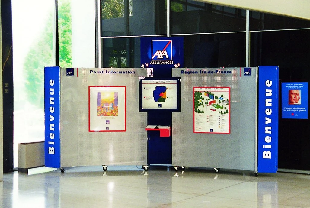 Stand d'accueil d'AXA Assurances dans un hall vitré : deux panneaux perforés « Point information » et « Région Île-de-France », bannières bleues « Bienvenue », écran au centre.