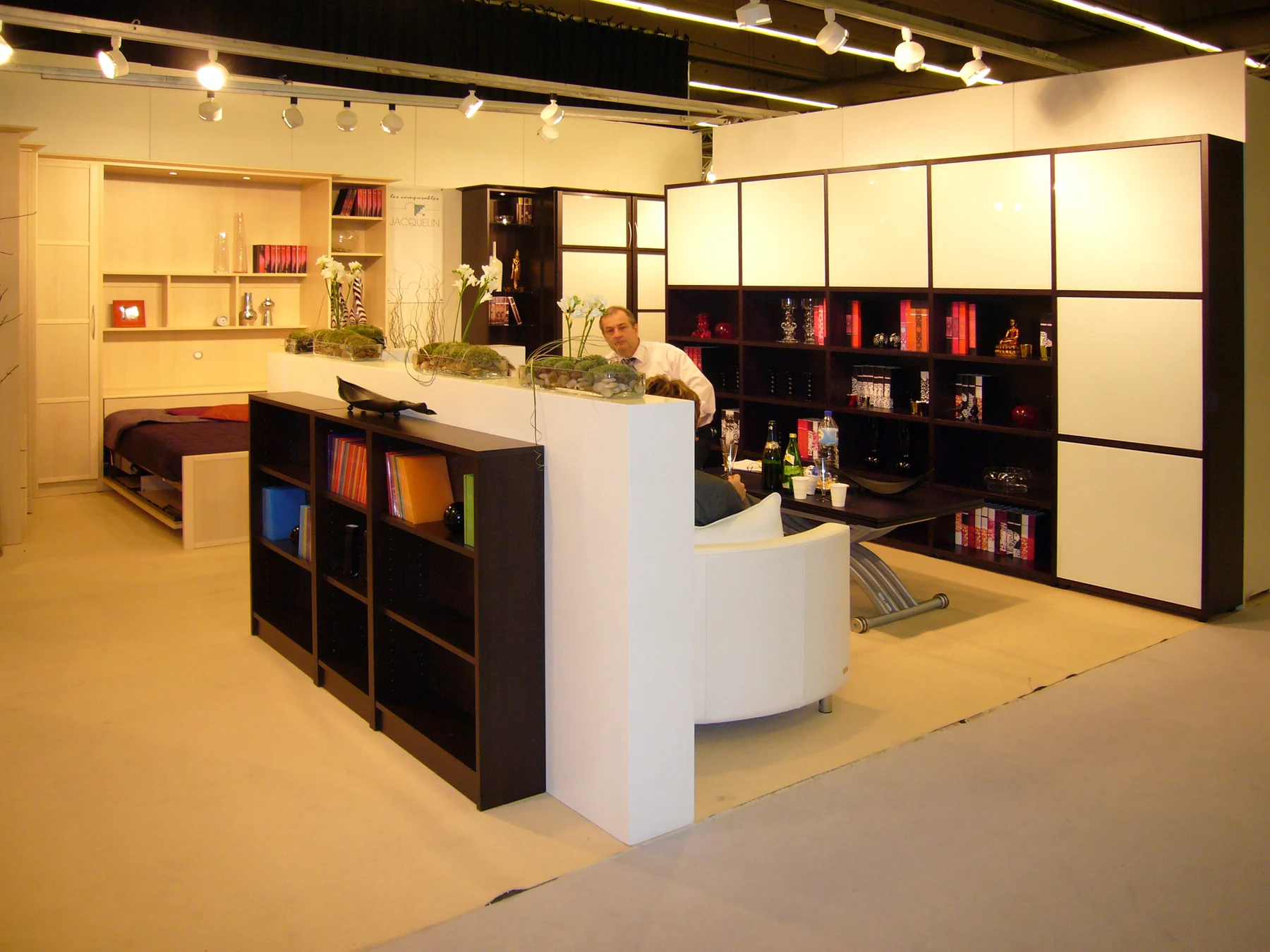 Stand des Composables Jacquelin au Salon du meuble : un îlot de librairies noires, un comptoir blanc et des ensembles meublés tout autour.