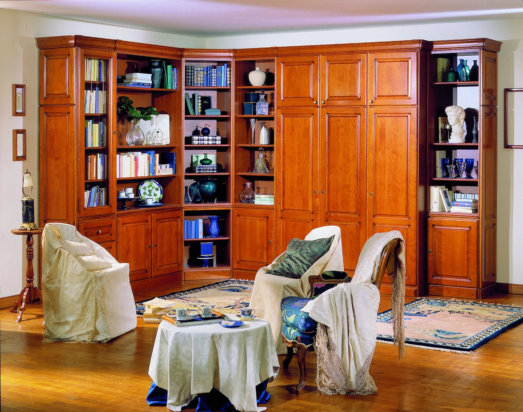 Salon en merisier : librairies et armoires du sol au plafond, fauteuils houssés de blanc, tapis.