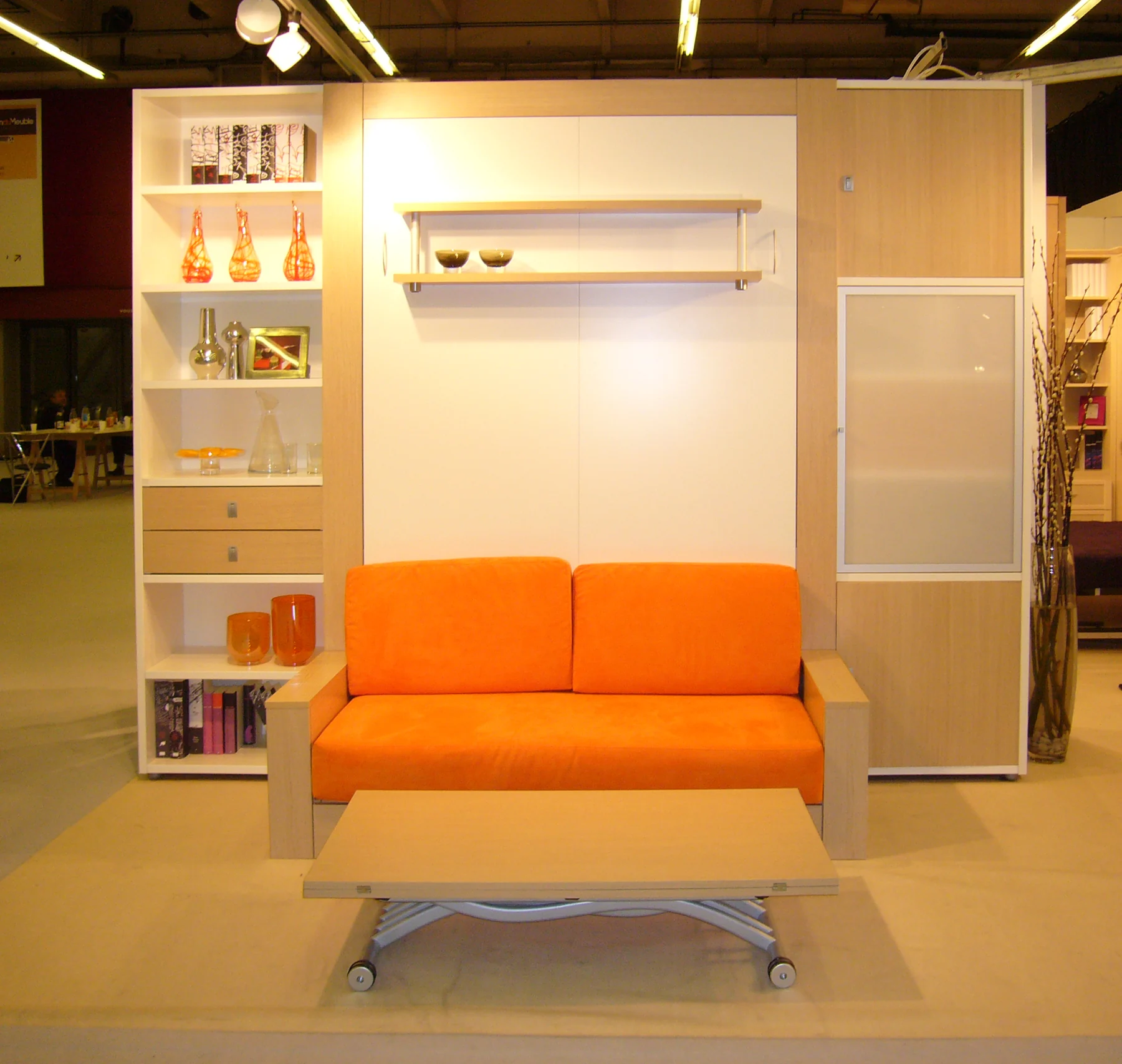 Ensemble clair sur le stand : librairies, lit escamoté derrière une façade blanche, canapé orange et table basse.
