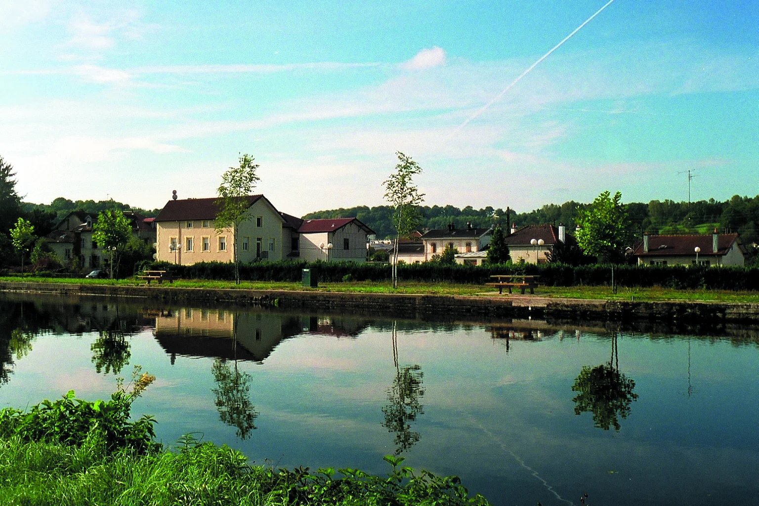 Le canal des Vosges en été, eau calme, maisons et arbres reflétés, ciel bleu.